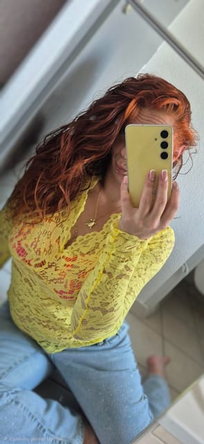 Selfie dans un miroir à l'extérieur, dans un escalier ensoleillé : une personne tient un smartphone devant elle et porte un haut en dentelle jaune et un jean bleu clair.
