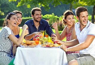 Sunny outdoor picnic with a group seated at a table filled with food and drinks, smiling under green trees in a warm, social atmosphere.