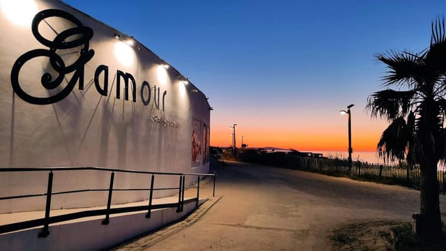 Vue extérieure du bâtiment du Glamour avec une grande enseigne, un chemin calme le long de la clôture et un ciel de coucher de soleil avec des palmiers à proximité à Cap d’Agde.
