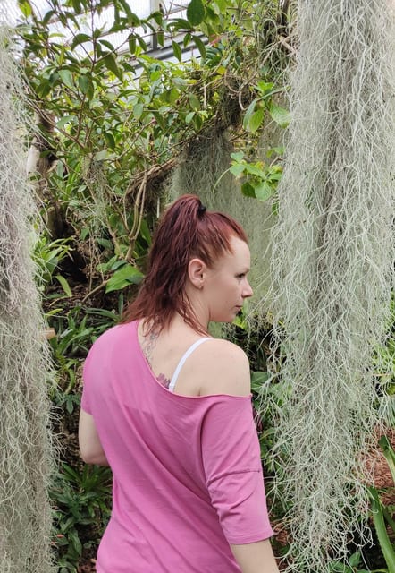 Personne en robe rose debout dans un jardin luxuriant, entourée de plantes vertes denses et de feuillage retombant, vue de profil.