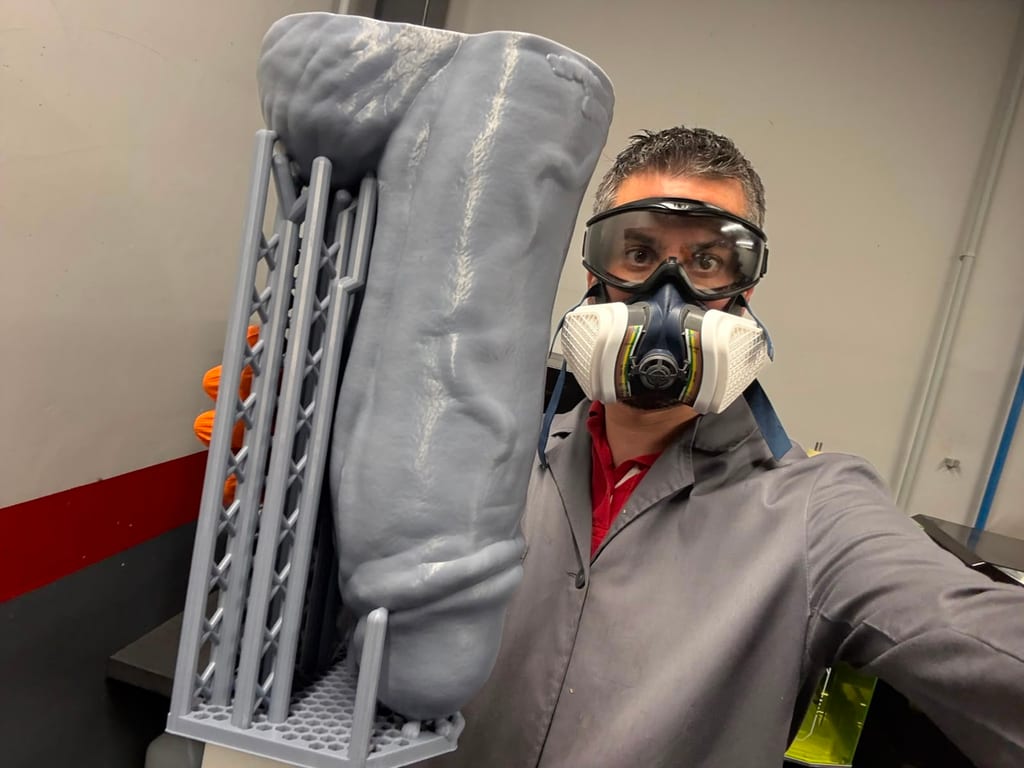Selfie dans un atelier : une personne porte des lunettes de protection et un masque respiratoire, près d’une grande pièce grise imprimée en 3D.