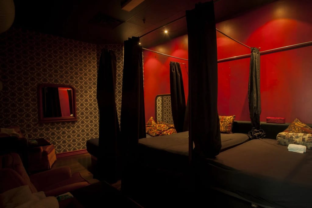 A red-accented room with patterned wallpaper, an illuminated mirror, and a curtained bed setup, lit with moody ambient light for @wishpride.
