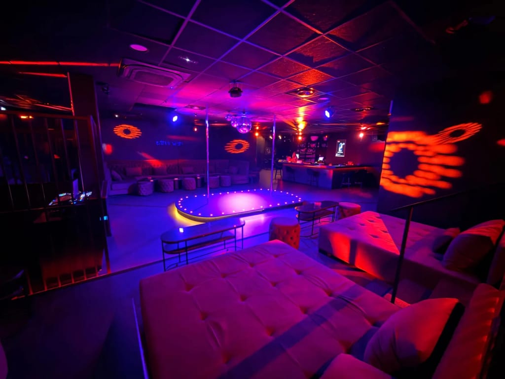 Image de couverture de @Bada Club montrant un lounge éclairé au néon, avec une plateforme circulaire lumineuse, des jeux de lumière colorés et des banquettes sous le plafond.