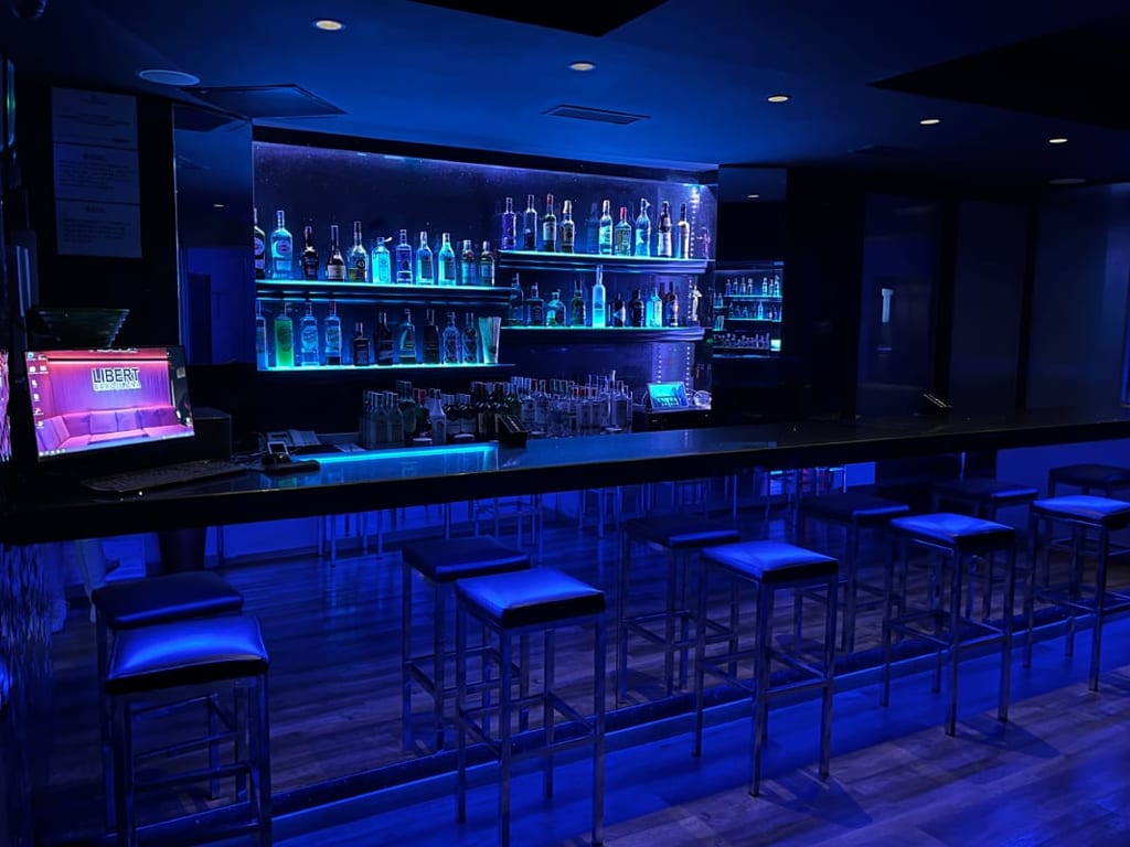Blue-lit bar interior with a long counter, shelves of bottles, and stools at Libert Swinger Club Barcelona in Barcelona.