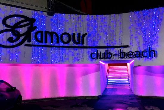 Couloir d’entrée du club de plage Le Glamour, éclairé en bleu et rose avec des guirlandes lumineuses scintillantes et un escalier menant à l’intérieur.