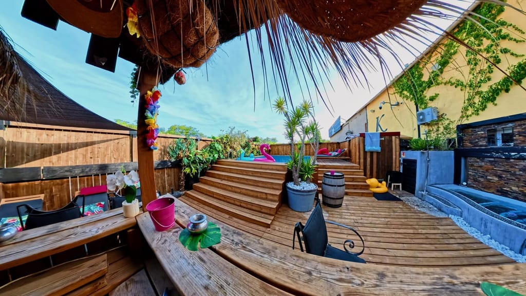 Terrasse extérieure en bois au style tropical chez Kalyptus by Rex, avec marches, plantes, décoration lounge colorée et un espace piscine ou bain à remous à droite.