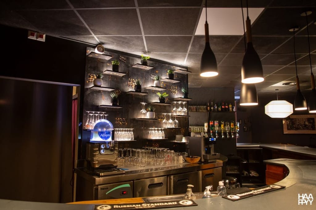 Intérieur du bar HAAPPYY avec des étagères de bouteilles et de verres, des suspensions lumineuses et un comptoir élégant dans une ambiance sombre et chaleureuse.