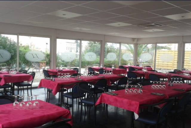 Salle à manger avec nappes rouges, assiettes et couverts soigneusement dressés, et grandes baies vitrées sous un plafond moderne et lumineux.