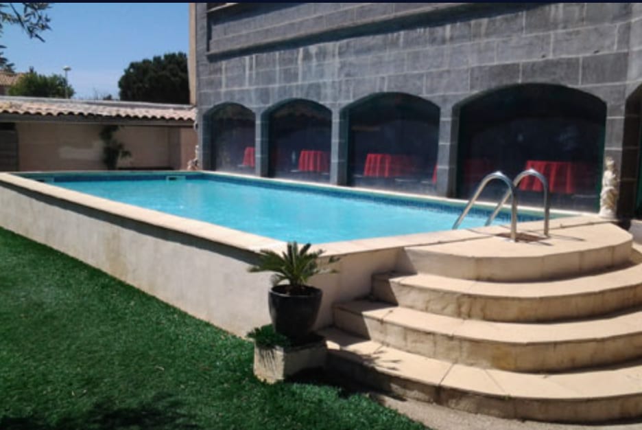 Piscine extérieure avec marches en pierre courbes, pelouse verte soigneusement entretenue et murs en pierre voûtés avec des chaises rouges à proximité.