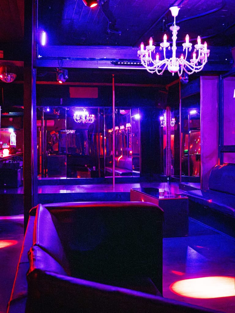 Photo de couverture de Cocoon Brussels montrant un lounge éclairé au néon, avec des lumières violettes et bleues, un lustre au plafond et un espace bar sombre.