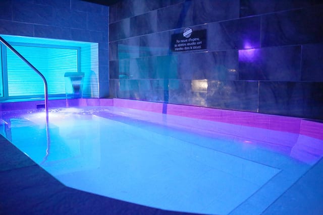Une piscine intérieure rectangulaire éclairée en bleu et violet se trouve dans une pièce carrelée, avec un panneau d'information au fond.