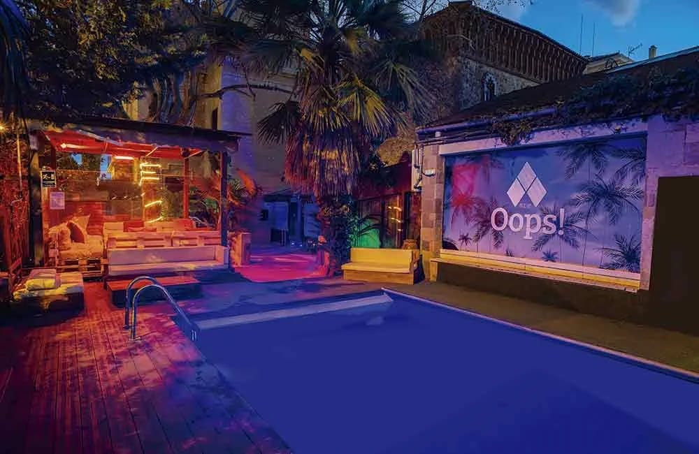 Neon-lit outdoor pool area with palm trees and modern building walls featuring the Oops! logo in Barcelona.