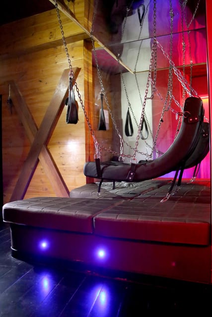 Intérieur éclairé au néon avec une chaise BDSM en cuir noir entourée de chaînes suspendues, sur fond de murs en bois et de contre-jours colorés.
