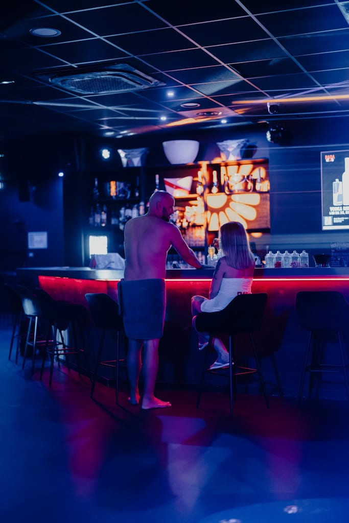 Intérieur du bar éclairé au néon du Bada Club, avec deux visiteurs assis sur des tabourets hauts, des cocktails et des bouteilles derrière le comptoir, sous une lumière d’ambiance bleu-violet.
