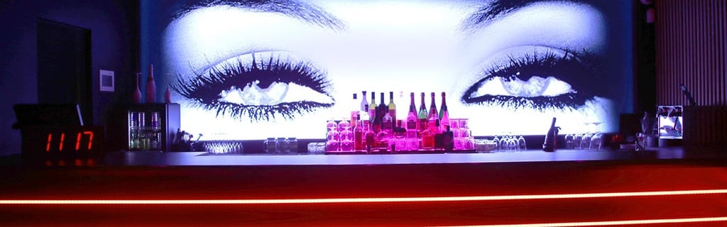 @Histoires d'Ô - Cap d'Agde cover montrant un comptoir de bar éclairé au néon avec des bouteilles colorées alignées derrière une bande rouge lumineuse.