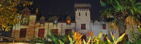 Bannière de @Château de Trédos montrant un château joliment éclairé au crépuscule, encadré par des arbres et des plantes colorées au premier plan.