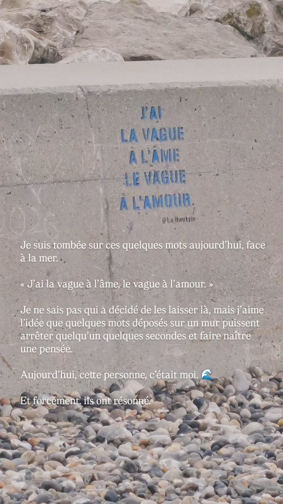 Scène en bord de mer depuis une plage de galets : un mur en béton avec une inscription bleue devant la mer, avec une citation superposée dans la publication @sarahwhisper.