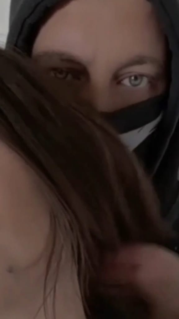 Portrait rapproché de @Eden_Coquin avec une capuche sombre et le visage couvert, seuls les yeux visibles, sur fond sombre à faible luminosité.