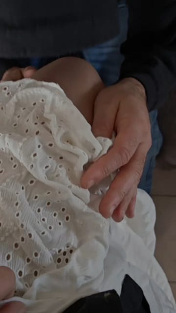 Gros plan sur des mains tenant doucement un tissu blanc à broderie anglaise, avec des vêtements en denim en arrière-plan, créant une ambiance chaleureuse et intime.
