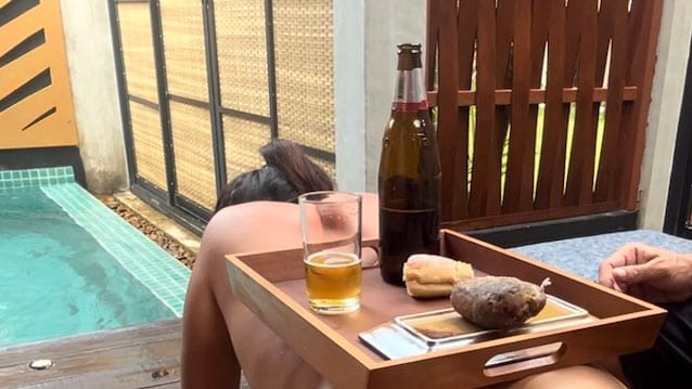 Un plateau de service en bois avec un verre de bière, une bouteille et du pain est posé près d’une piscine turquoise et d’un paravent en bois.