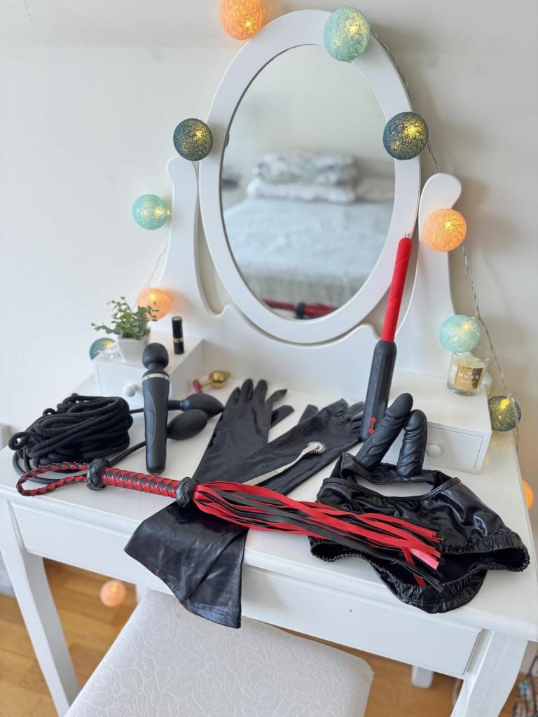 Mise en scène à thème BDSM sur une coiffeuse blanche : un miroir ovale entouré de guirlandes lumineuses, des gants en satin noir et des accessoires rouge et noir soigneusement disposés.